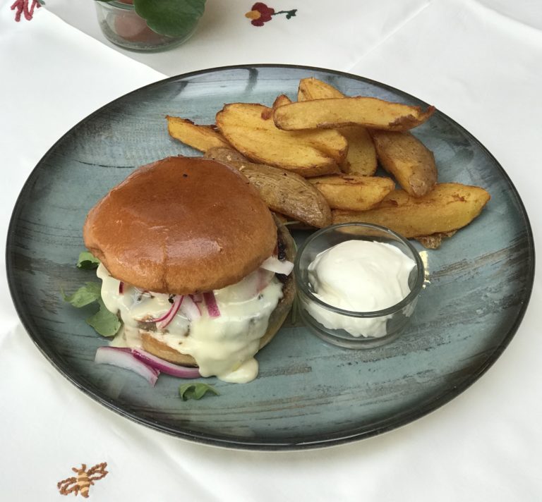 Chaussee-Burger auf einem türkisen Teller, mit selbstgemachtem Kochkäse, selbstgemachter Barbecuesoße, Jalapeños, Zwiebeln und Salat, daneben ein Schälchen Sour Cream und Kartoffelspalten